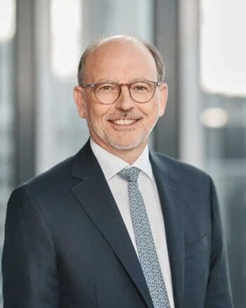 Das Bild zeigt Thomas Groß, CEO der Helaba, in einem formellen Anzug mit einer Krawatte. Er lächelt freundlich und steht vor einer modernen, lichtdurchfluteten Umgebung. Die Aufnahme vermittelt Professionalität und Offenheit. Im Kontext der Teilnahme an den EZB-Trials für Distributed Ledger Technologie-Lösungen unterstreicht Thomas Groß die Bedeutung des Wissensaustauschs und der Stärkung der Rolle der Helaba in zukünftigen digitalen Lösungen.