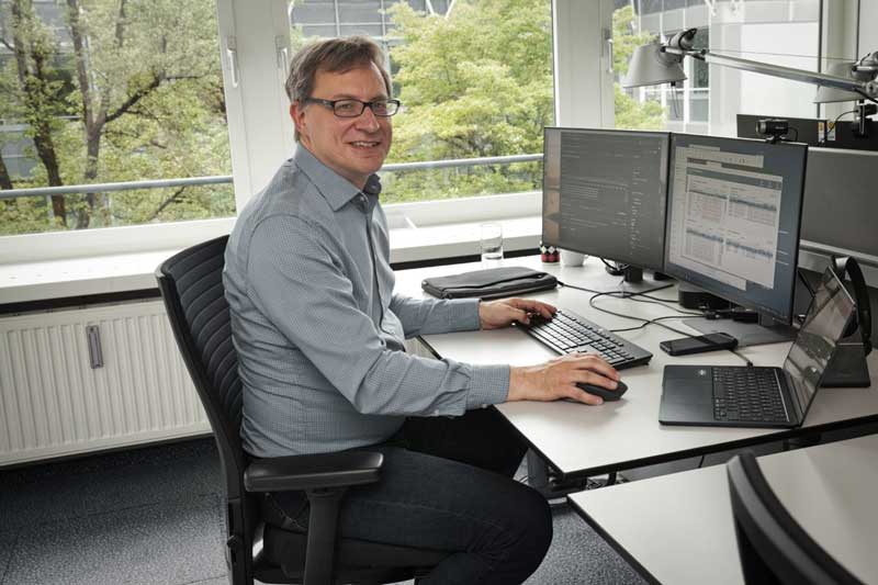 Das Bild zeigt Christian Brunner, der im Büro von FI-TS in Haar bei München an einem Schreibtisch sitzt. Er arbeitet an mehreren Monitoren, die Informationen zu Cloud Native-Anwendungen anzeigen.
