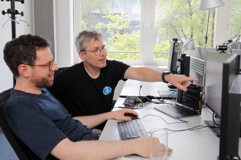 Das Bild zeigt zwei Personen, die an einem Schreibtisch sitzen und auf einen Computerbildschirm schauen. Der Mann links, Stefan Majer, und der Mann rechts, Gerrit Schwerthelm, diskutieren über technische Details. Auf dem Tisch befinden sich mehrere Computer und Zubehör. Die Umgebung ist hell und modern, mit großen Fenstern, die Tageslicht hereinlassen. Diese Szene verdeutlicht die Zusammenarbeit im Rahmen der Entwicklung einer Cloud Native Infrastruktur.