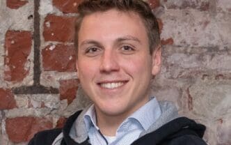 Das Bild zeigt einen jungen Mann, der freundlich in die Kamera lächelt. Er trägt ein hellblaues, kariertes Hemd und einen dunklen Kapuzenpullover, was ihm einen lässigen Look verleiht. Der Hintergrund besteht aus einer sichtbaren Backsteinwand mit roten und grauen Steinen, die dem Bild einen rustikalen Charme verleihen. Die Beleuchtung ist weich und sorgt für eine angenehme Atmosphäre. Der Kontext deutet auf eine entspannte Umgebung hin, möglicherweise in einem Café oder einem kreativen Arbeitsraum.