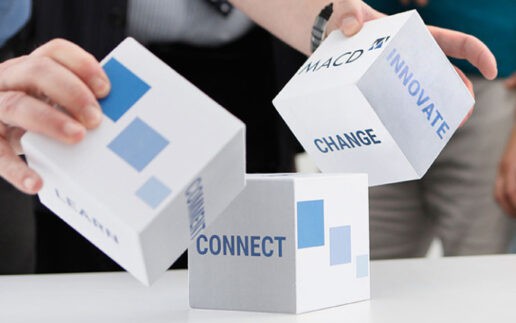 Auf dem Bild sind mehrere Würfel zu sehen, die in der Luft schweben. Die Würfel tragen die Begriffe „CONNECT“, „INNOVATE“, „CHANGE“ und „LEARN“. Eine Person hält einen Würfel, während im Hintergrund weitere Personen sichtbar sind. Der Kontext deutet auf eine kreative oder strategische Diskussion hin.