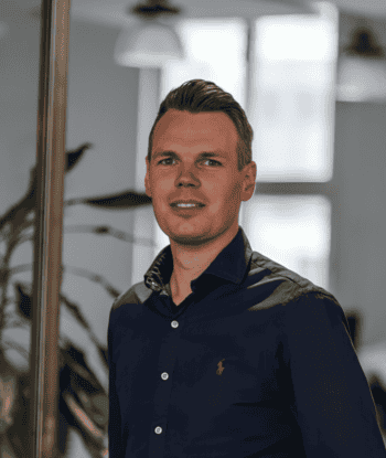 Moritz Gerdes, Head of signals, dem Innovations-Ökosystem der SIGNAL IDUNA Gruppe zum Innovations-Management, ist in einem modernen Büro abgebildet. Er trägt ein dunkelblaues Hemd und steht vor einer Glaswand, die einen Blick auf die Büroeinrichtung ermöglicht. Die Umgebung ist hell und freundlich gestaltet, was auf eine innovative Arbeitsatmosphäre hinweist. Gerdes repräsentiert die strategische Ausrichtung des Unternehmens im Bereich Zukunftstechnologien und Innovationskultur.