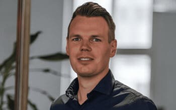 Das Bild zeigt Moritz Gerdes, Head of signals der SIGNAL IDUNA Gruppe. Er präsentiert sich in einem professionellen Umfeld, umgeben von Pflanzen, was auf eine moderne Arbeitsatmosphäre hinweist. Gerdes ist verantwortlich für das Innovations-Management innerhalb des Unternehmens und agiert als Brückenbauer zwischen verschiedenen Akteuren im Innovationsökosystem. Sein Auftreten vermittelt Kompetenz und Engagement für technologische Trends und Entwicklungen.