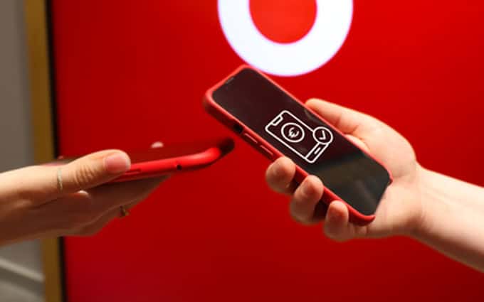 Das Bild zeigt zwei Hände, die jeweils ein rotes Smartphone halten. Die linke Hand gehört einer Person, die ein Smartphone mit einem klaren Display zeigt, auf dem ein Symbol für eine Geldtransaktion abgebildet ist. In der rechten Hand wird ebenfalls ein rotes Smartphone präsentiert. Der Hintergrund ist in einem kräftigen Rot gehalten und zeigt ein Logo von Vodafone, was auf einen Telekommunikationskontext hinweist. Die Darstellung vermittelt den Eindruck einer modernen Zahlungstechnologie, die möglicherweise in einem Geschäft oder einer Verkaufsstelle genutzt wird.