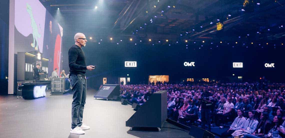 Scott Galloway steht auf der Bühne der OMR 2025 und präsentiert seinen jährlichen Vortrag. Er trägt einen schwarzen Pullover und Jeans, während er ein Publikum von mehreren Hundert Zuhörern anspricht. Im Hintergrund sind große Bildschirme mit grafischen Darstellungen zu sehen. Die Atmosphäre ist geprägt von einer professionellen und aufmerksamen Stimmung, die den traditionellen Auftakt zur OMR-Veranstaltung unterstreicht.