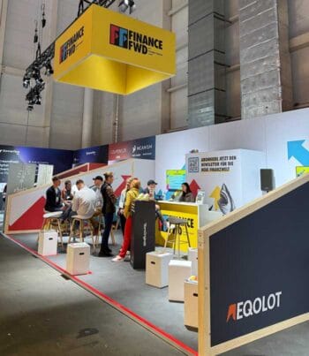 Das Bild zeigt den Stand von Finance Forward (FFWD) auf dem OMR-Festival in Halle 4. Der Stand ist modern gestaltet und umfasst mehrere Sitzgelegenheiten sowie Informationsstände. Besucher interagieren miteinander, während Mitarbeiter Informationen bereitstellen. Im Hintergrund sind weitere Marken und Sponsoren sichtbar, die das Event unterstützen. Die Atmosphäre wirkt geschäftig und einladend, was auf das Interesse an den Themen der Finanzwelt hinweist.