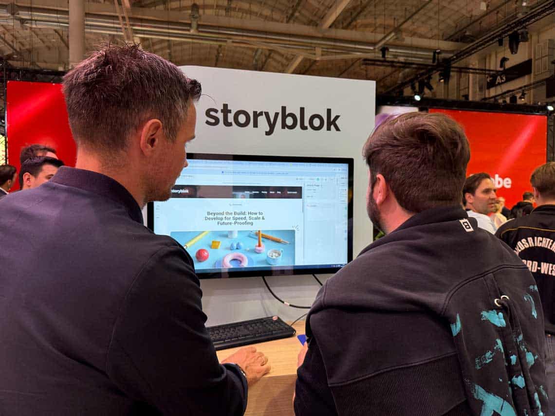 Das Bild zeigt zwei Personen, die an einem Computerbildschirm sitzen, auf dem eine Webseite von Storyblok angezeigt wird. Die Umgebung ist geprägt von einem Messe-Setting, erkennbar an den Ständen im Hintergrund. Die Veranstaltung findet im Rahmen des OMR-Events statt, das sich mit digitalen Trends und Innovationen beschäftigt. Die Personen sind in lockerer Kleidung gekleidet und scheinen sich auf die Inhalte des Bildschirms zu konzentrieren.
