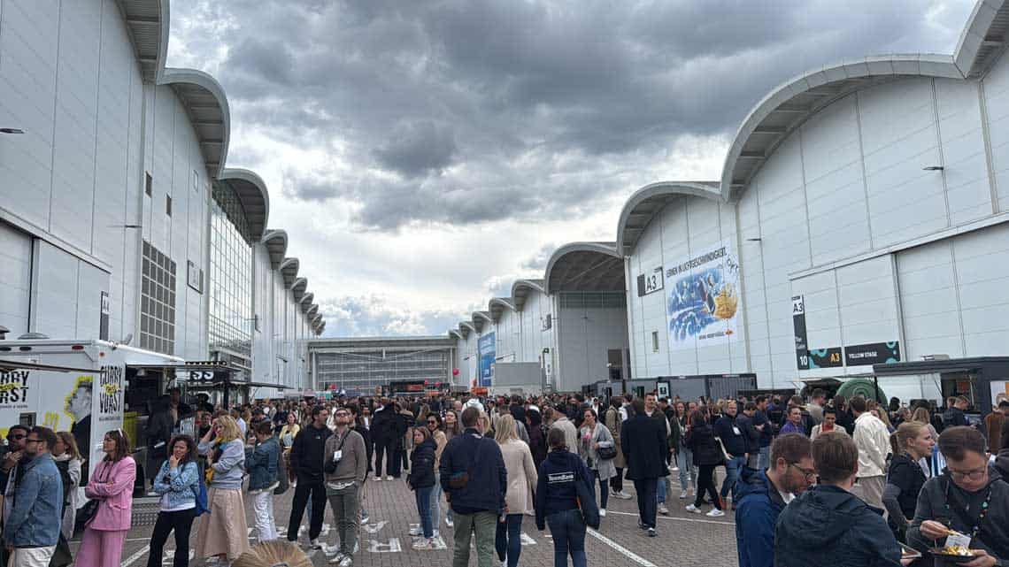 Das Bild zeigt eine große Menschenmenge, die sich vor einer modernen Messehalle versammelt hat. Die Teilnehmer scheinen sich in Gesprächen zu befinden und verschiedene Essensstände zu besuchen. Über den Köpfen der Menschen sind graue Wolken sichtbar, die eine düstere Stimmung vermitteln. Die Veranstaltung steht im Kontext von OMR, wo zahlreiche Masterclasses angeboten wurden, die den Besuchern wertvolle Inhalte und Networking-Möglichkeiten boten.
