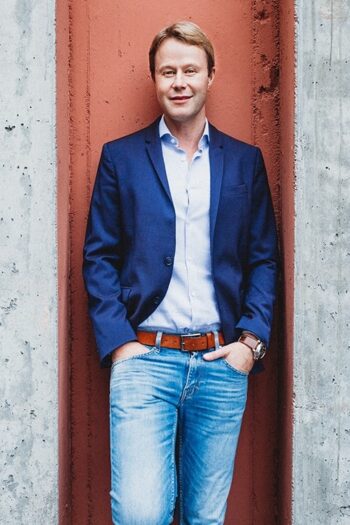 Ein Mann in formeller Kleidung steht entspannt vor einer Wand mit rotem Putz. Er trägt ein blaues Sakko und Jeans. Diese Darstellung könnte mit der Kooperation von Iwoca und Smava in Verbindung stehen, die alternative Finanzierungslösungen für KMU anbieten.