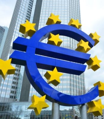 Das Bild zeigt das markante Euro-Symbol, umgeben von gelben Sternen, vor einem modernen Hochhaus. Diese ikonische Darstellung steht symbolisch für die Europäische Zentralbank (EZB) und deren Rolle im europäischen Finanzsystem. Die Architektur im Hintergrund unterstreicht den urbanen Charakter des Standorts, der für Innovationen im Zahlungsverkehr, wie den digitalen Euro, von Bedeutung ist. Die Farben Blau und Gelb dominieren das Bild und repräsentieren die europäische Identität.