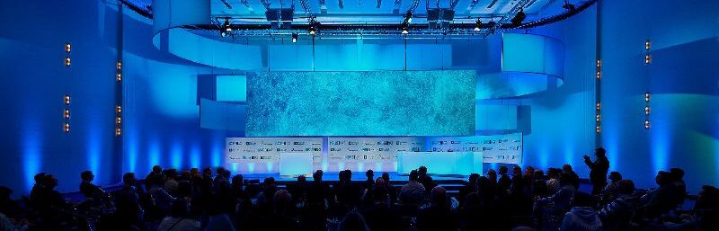 Das Bild zeigt den Veranstaltungsraum der Kongressmesse InsureNXT 2025 in Köln. Die Bühne ist mit einem großen, beleuchteten Bildschirm ausgestattet, der in sanften Blautönen gehalten ist. Im Vordergrund sind zahlreiche Teilnehmer zu sehen, die auf die Präsentation blicken. Die Anordnung der Sitzplätze und die moderne Gestaltung des Raumes unterstreichen die Innovationsorientierung der Veranstaltung, die sich auf digitale Transformation und künstliche Intelligenz in der Versicherungswirtschaft konzentriert.