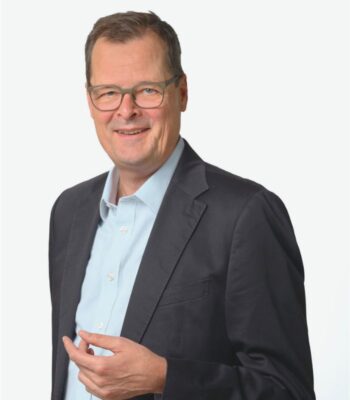 Das Bild zeigt einen Mann in einem dunklen Blazer und hellblauen Hemd, der freundlich in die Kamera lächelt. Er steht mit einer Hand leicht erhoben, was auf eine offene und kommunikative Haltung hinweist. In der Rolle als Innovationspartner der EZB wird er zur Entwicklung des digitalen Euro beitragen, um dessen Mehrwerte für die Wirtschaft zu fördern. Der Hintergrund ist neutral gehalten, was den Fokus auf die Person lenkt.