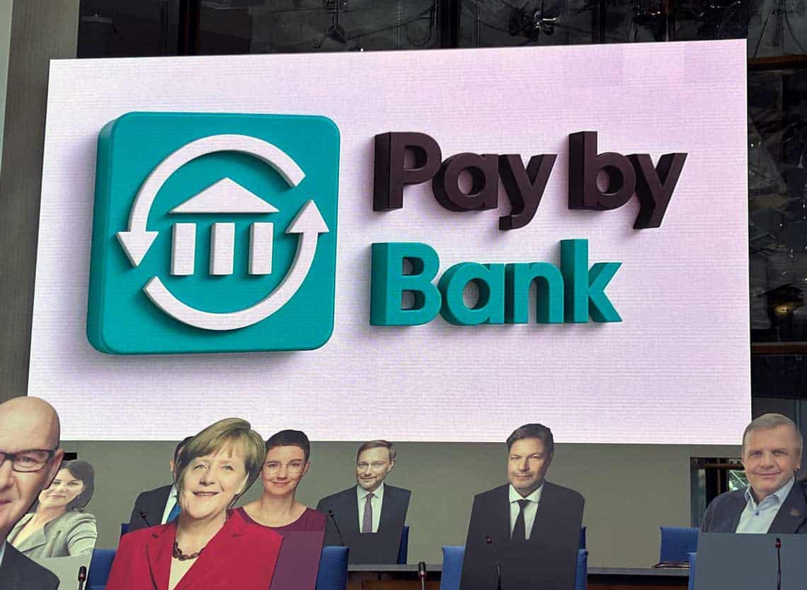 Das Bild zeigt ein großes, digitales Banner mit dem Schriftzug „Pay by Bank“ in auffälligen Farben - das neue Pay-by-Bank-Logo. Im Hintergrund sind mehrere Pappaufsteller von beteiligten Politikern. Die Darstellung des Logos symbolisiert eine neue Ära im Zahlungsverkehr, die durch innovative Verfahren wie „Pay by Bank“ geprägt ist.