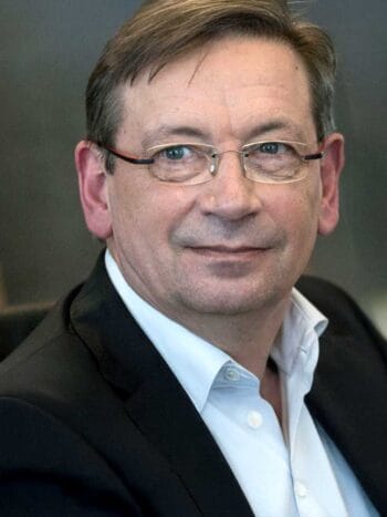 Das Bild zeigt Dr. Rainer Baumgart, Co-Founder von eleQtron. Er trägt einen dunklen Anzug mit einem weißen Hemd. Die Präsentation von Dr. Baumgart steht im Kontext der Diskussion über Quantencomputer, die potenziell in der Lage sind, komplexe Rechenprobleme in der Finanz- und Sicherheitsbranche effizient zu lösen.