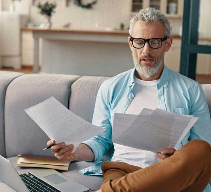 Ein Mann mit Brille sitzt auf einem Sofa und betrachtet mehrere Dokumente. Vor ihm liegt ein Laptop. Die Umgebung ist hell und modern gestaltet. Der Fokus liegt auf der Analyse von Informationen, die möglicherweise im Kontext von DORA relevant sind.