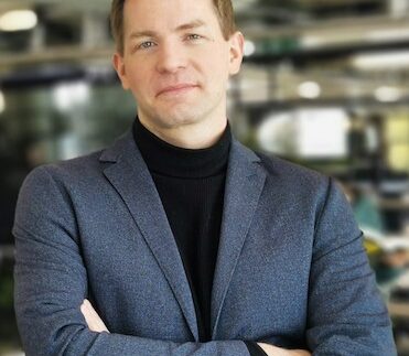 Ein Mann in einem grauen Blazer und schwarzem Rollkragenpullover steht mit verschränkten Armen in einem modernen Büro. Die Umgebung deutet auf eine innovative Arbeitsatmosphäre hin. Diese Kooperation zwischen Iwoca und smava adressiert den Finanzierungsbedarf von KMU.
