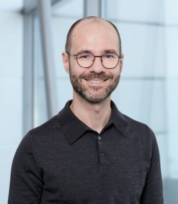Das Bild zeigt Oliver Dörler, der als Chief Data & AI Officer der Commerzbank tätig ist. Er trägt ein schwarzes Polohemd und hat eine Brille. Im Hintergrund sind moderne architektonische Elemente zu erkennen, die eine helle und transparente Atmosphäre schaffen. Dörler lächelt freundlich in die Kamera, was eine positive Ausstrahlung vermittelt.