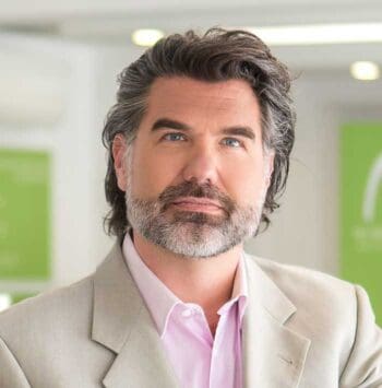 Ein Mann mit grauen Haaren und Bart trägt einen hellen Anzug und ein rosa Hemd. Der Hintergrund ist unscharf, mit grünen Elementen, die auf eine moderne Umgebung hinweisen. Der Fokus liegt auf dem Thema Raster und dessen Einfluss auf die Versicherungs-IT.