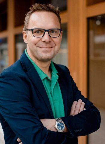 Christian Berger, Geschäftsführer von Finnoconsult, präsentiert sich in einem professionellen Umfeld. Er trägt einen blauen Blazer und ein grünes Polohemd, während er mit verschränkten Armen lächelt. Der Hintergrund deutet auf eine moderne Architektur hin.
