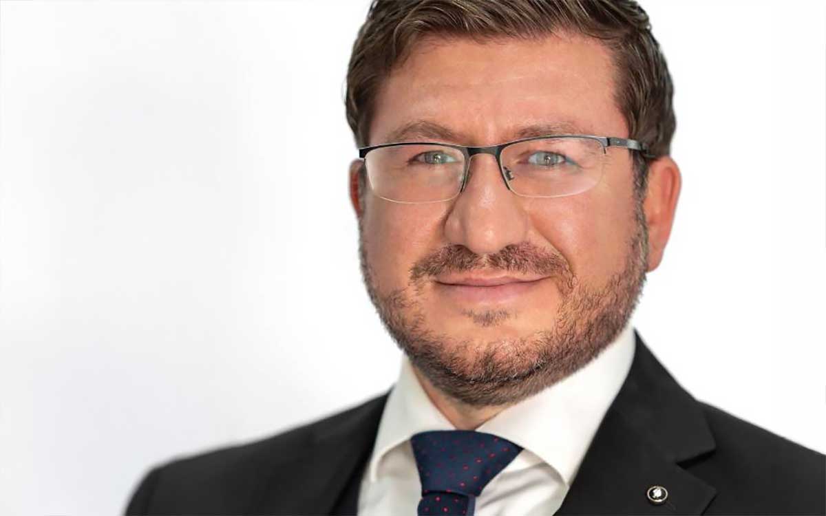 Ein Mann mit Brille und gepflegtem Bart trägt einen Anzug mit Krawatte. Der Hintergrund ist neutral und hell. Die Darstellung vermittelt Professionalität und könnte im Kontext von Blackout-Strategien in der Finanzbranche verwendet werden, um Mitarbeiter-Awareness zu fördern.