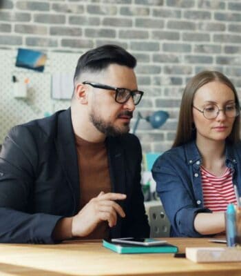 Zwei Personen arbeiten an einem Tisch in einem modernen Büro. Der Mann mit Brille und Anzug diskutiert intensiv mit der Frau, die eine gestreifte Bluse trägt. Im Hintergrund sind Büromaterialien sichtbar. Die Kooperation zwischen S-Payment und ePages wird thematisiert.
