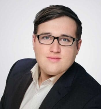 Ein junger Mann mit Brille und einem dunklen Anzug blickt direkt in die Kamera. Sein Gesichtsausdruck ist neutral. Die Anforderungen an Compliance im IT-Outsourcing sind entscheidend, um Kontrollverlust und mangelnde Nachweisfähigkeit zu vermeiden.