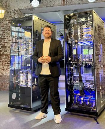Ein Mann steht zwischen zwei IBM Mainframes in einem modernen Veranstaltungsraum. Die Geräte sind transparent und beleuchtet, was ihre technische Komplexität betont. Diese Mainframes gelten als das IT-Rückgrat vieler Unternehmen und unterstützen essentielle Kernprozesse.