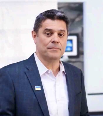 Kerim Akgonul, CPO bei Pegasystems, ist in einem modernen Büro abgebildet. Er trägt einen Anzug und hat ein Emblem an seinem Revers. Im Hintergrund sind digitale Displays zu erkennen, die auf technologische Innovationen hinweisen.