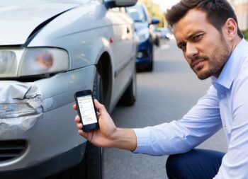Ein Mann kniet neben einem beschädigten Auto und hält ein Smartphone in der Hand. Auf dem Bildschirm ist eine Anwendung zur Schadenabwicklung sichtbar. Die Szene verdeutlicht die zunehmende digitale Interaktion bei der Schadensregulierung.