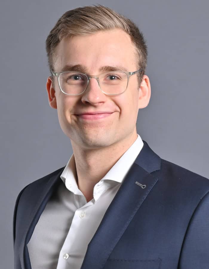 Tim Zölitz, CEO von Crypto Risk Metrics, ist hier mit Brille und dunkelblauem Anzug vor grauem Hintergrund abgelichtet.