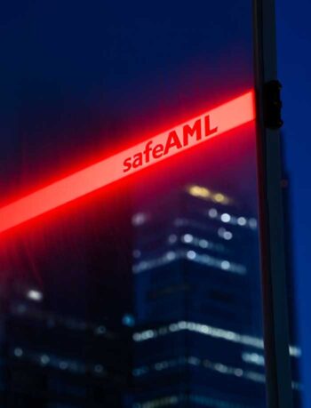 Ein rotes Lichtband mit der Aufschrift „safeAML“ ist vor einer urbanen Nachtlandschaft positioniert. Die unscharfen Konturen von Hochhäusern im Hintergrund deuten auf eine städtische Umgebung hin, die mit Finanzaktivitäten assoziiert wird.