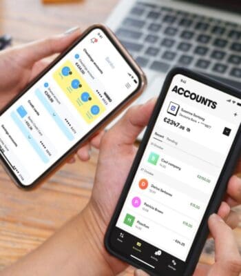 Zwei Smartphones werden gehalten, auf denen digitale Wallets angezeigt werden. Eine der Apps zeigt die Funktionalität von Wero, während die andere Kontoinformationen präsentiert. Die Geräte sind auf einem Holztisch platziert, im Hintergrund ist ein Laptop sichtbar.