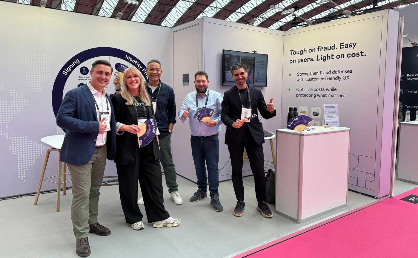 Fünf Personen stehen an einem Messestand bei Money20/20. Sie halten Fächer in der Hand und posieren freundlich für die Kamera. Im Hintergrund sind Informationen zu Betrugsprävention und Benutzerfreundlichkeit sichtbar. Der Stand vermittelt ein modernes, professionelles Ambiente.