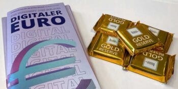 Ein Buch mit dem Titel „Digitaler Euro“ liegt auf einem Tisch, umgeben von goldenen Schokoladentafeln, die mit „GOLD RESERVE“ beschriftet sind. Die Darstellung thematisiert die Diskussion um den digitalen Euro als Infrastrukturprojekt und Konsumentenprodukt.