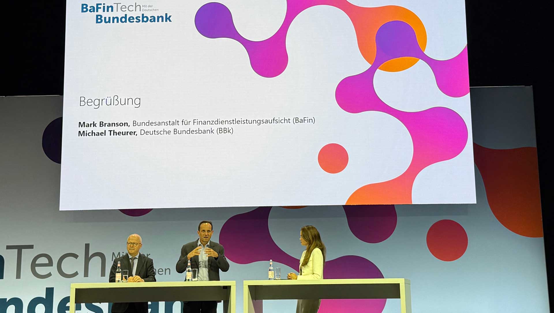 Mark Branson, Präsident der BaFin, und Michael Theurer, Vorstand der Deutschen Bundesbank, stehen an einem Tisch und diskutieren auf der BaFinTech 2025. Im Hintergrund ist eine grafische Darstellung mit bunten Elementen zu sehen.