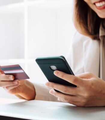 Eine Person hält ein Smartphone in der rechten Hand und eine Kreditkarte in der linken Hand. Im Hintergrund ist ein Laptop sichtbar. Diese Darstellung könnte im Kontext der neuen „easyCredit-Rechnung“ von TeamBank im E-Commerce stehen.