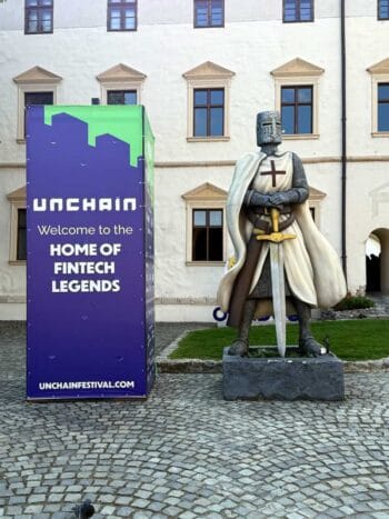 Ein Werbeschild des UNCHAIN Festivals steht neben einer Ritterstatue in Oradea. Das Schild verkündet „Welcome to the HOME OF FINTECH LEGENDS“. Im Hintergrund sind historische Gebäude mit Fenstern sichtbar.