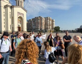 Teilnehmende des UNCHAIN Festivals versammeln sich in der Altstadt von Oradea. Eine Person erklärt Informationen, während die Gruppe aufmerksam zuhört. Im Hintergrund sind moderne Gebäude und eine Kirche sichtbar, die die historische Kulisse ergänzen.
