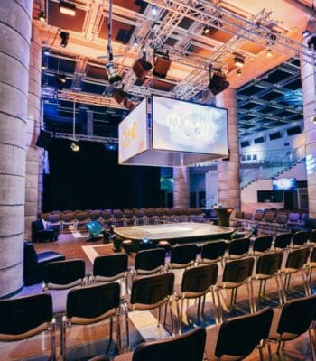 Die Event-Location Pyramide Mainz ist für die Veranstaltung „PayForward“ vorbereitet. Der Raum ist mit Stühlen ausgestattet, und eine zentrale Bühne ist sichtbar. Beleuchtung und audiovisuelle Technik sind installiert, um eine ansprechende Atmosphäre zu schaffen.