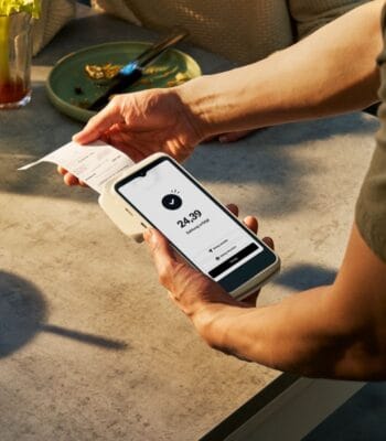 Ein Mobilgerät zeigt einen Betrag von 24,39 an, während eine Person einen Beleg übergibt. Die Szene vermittelt die Nutzung eines Mobile POS zur Zahlungsabwicklung in einem gastronomischen Umfeld. Ein Teller und Besteck sind im Hintergrund sichtbar.