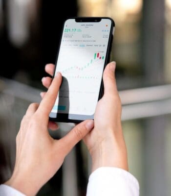 Eine Person nutzt ein Smartphone, um eine Handelsanwendung zu bedienen. Auf dem Bildschirm sind Aktienkurse und Diagramme zu sehen, was auf das wachsende Interesse an Neobrokern hinweist. Die Umgebung ist modern und hell.