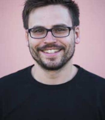 Patrick Ring, neuer Head of Data Analytics & KI beim Münchener Verein, lächelt in die Kamera. Er trägt ein schwarzes T-Shirt und hat eine Brille auf. Der Hintergrund ist in einem sanften Rosa gehalten.