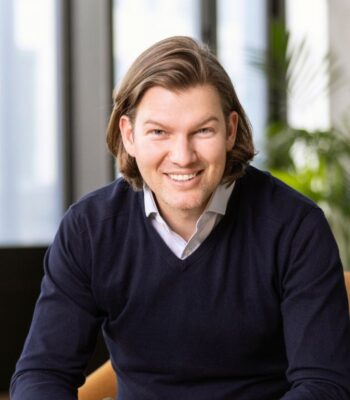 Valentin Stalf, Gründer und Co-CEO von N26, lächelt in einem modernen Büro. Er trägt einen dunkelblauen Pullover und sitzt entspannt auf einem Stuhl. Im Hintergrund sind Pflanzen und große Fenster sichtbar, die Tageslicht hereinlassen.