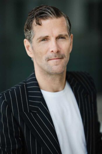 Patric Dahse mit gestreiftem Blazer und T-Shirt blickt direkt in die Kamera. Sein Gesichtsausdruck vermittelt Selbstbewusstsein. Die Abbildung könnte im Kontext der Diskussion über Technologie und IT-Transformation in der Finanzbranche stehen.