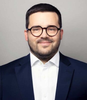 Philip Mayer, Senior Manager bei Cofinpro, präsentiert sich in einem formellen Outfit. Sein Lächeln und die Brille verleihen ihm ein professionelles Erscheinungsbild. Im Kontext des Wertpapiergeschäfts symbolisiert er die Verbindung zwischen Tradition und digitaler Innovation.