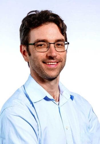 Ein Mann mit Brille und einem lächelnden Gesicht trägt ein hellblaues Hemd. Er steht vor einem einfarbigen Hintergrund. In der Versicherungsbranche könnte Agentic AI eine transformative Rolle spielen, indem sie proaktive und autonome Lösungen bietet.