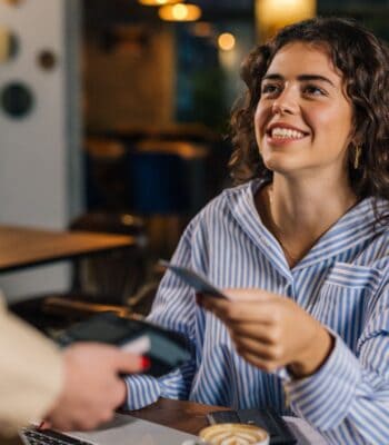 Eine junge Frau lächelt, während sie eine girocard in der Hand hält und diese einem Service-Mitarbeiter überreicht. Im Hintergrund sind Tische und eine gemütliche Café-Atmosphäre zu erkennen.