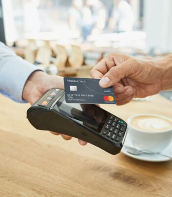 Eine Hand hält eine Mastercard, während sie über ein Kartenlesegerät gehalten wird. Im Hintergrund sind unscharfe Personen und eine Tasse Kaffee sichtbar. Die Transaktion symbolisiert moderne Zahlungstechnologien im Zahlungsverkehr.