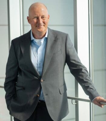 Michael Rasch, neuer Vorstand bei msg, steht lächelnd an einem Geländer in einem modernen Bürogebäude. Er trägt einen grauen Anzug und ein hellblaues Hemd, was seine Professionalität unterstreicht. Der Hintergrund ist hell und minimalistisch gestaltet.