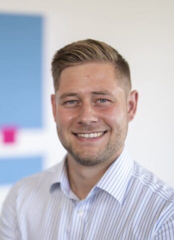 Steffen Grenzheuser, DPS, lächelt in die Kamera. Der Hintergrund ist hell und modern gestaltet, mit blauen und pinken Elementen. Die Darstellung vermittelt Professionalität und Offenheit, was im Kontext der Diskussion um ISO 20022 relevant ist.